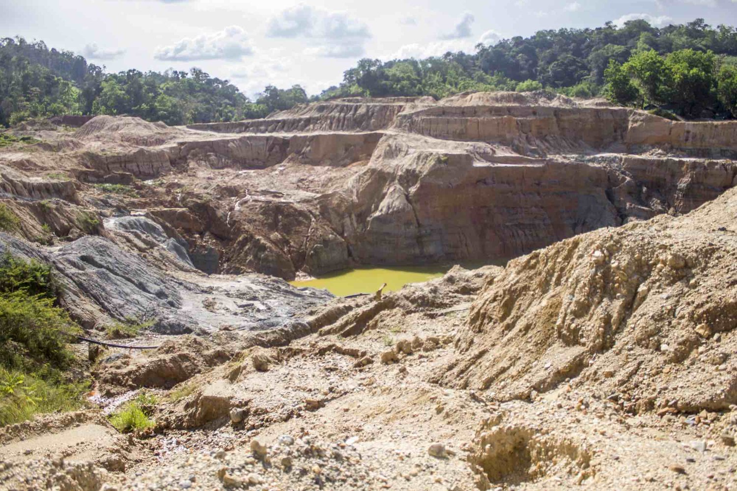 The illegally mined land is a shockingly barren moonscape… massive mountains of bare, eroding orange soil linked to one another by a chain of stagnant red-brown to neon-green mine waste ponds, in ugly contrast to the surrounding verdant, bio-diverse and life-giving jungle.